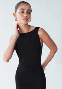 Black sleeveless dress with a round neckline, smooth texture, fitted silhouette, worn with gold hoop earrings and a chunky bracelet.