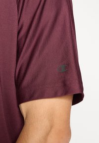 Bordeaux kortemouwshirt van zacht materiaal, met een ronde hals en een subtiel logo op de linkermouw. Soepele textuur, casual ontwerp.