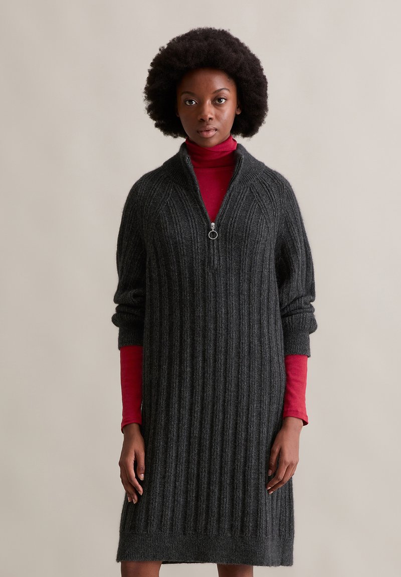 Donkergrijze geribde jurk met een kwart-rits kraag en een relaxte pasvorm. Lange mouwen met een subtiele manchetafwerking. Gelaagd over een rode coltrui.