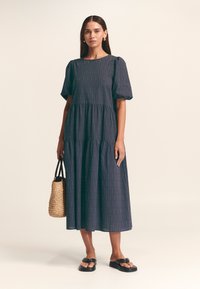 Abito blu navy a balze con maniche a palloncino corte e motivi geometrici dorati. Il modello tiene in mano una borsa intrecciata e indossa sandali neri flat.