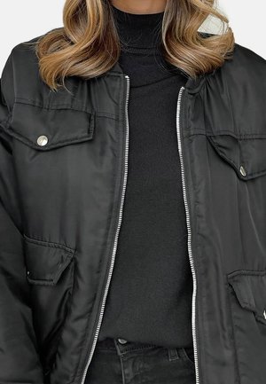 Femme portant un blouson bomber noir avec fermeture éclair argentée et poches à boutons-pression sur un pull à col roulé noir.