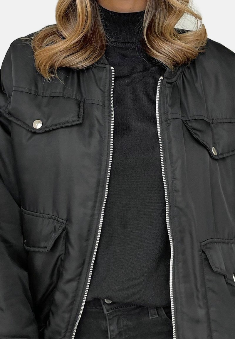 Femme portant un blouson bomber noir avec fermeture éclair argentée et poches à boutons-pression sur un pull à col roulé noir.