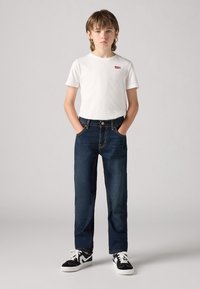 Bijela pamučna majica s logom na prsima, u kombinaciji s tamnoplavim trapericama i crno-bijelim tenisicama. Ravnog kroja.