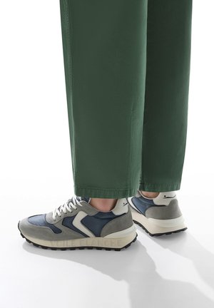 Sneaker mit grauem und marineblauem Obermaterial, weißen Akzenten und einer klobigen cremefarbenen Zwischensohle. Verfügt über strukturiertes Wildleder und eine Gummi-Außensohle. Grüne Hose ist sichtbar.