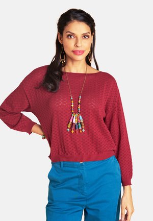 Frau mit langem dunklem Haar trägt einen strukturierten roten Pullover, blaue Hose, eine bunte Perlenkette und passende Ohrringe, steht mit einer Hand in der Hüfte.