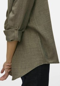 Olijfgroene shirt met een gestructureerde stof, opgerolde mouw en een gebogen zoom. Heeft een knoopdetail op de mouw en een rechte pasvorm.