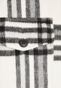 Tela de cuadro en blanco y negro con una textura suave. Presenta un bolsillo con un botón negro y un detalle de costura en contraste.