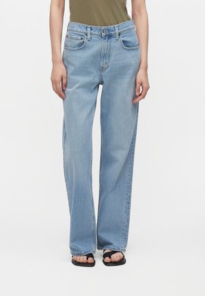 Personne portant un jean large bleu clair, un haut vert olive et des sandales noires, debout devant un fond blanc uni.