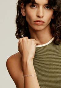 Pulsera de plata con acentos de estrellas llevada en una mano. La blusa de canalé verde del modelo presenta un ribete blanco a lo largo del escote. Textura de piel suave visible.