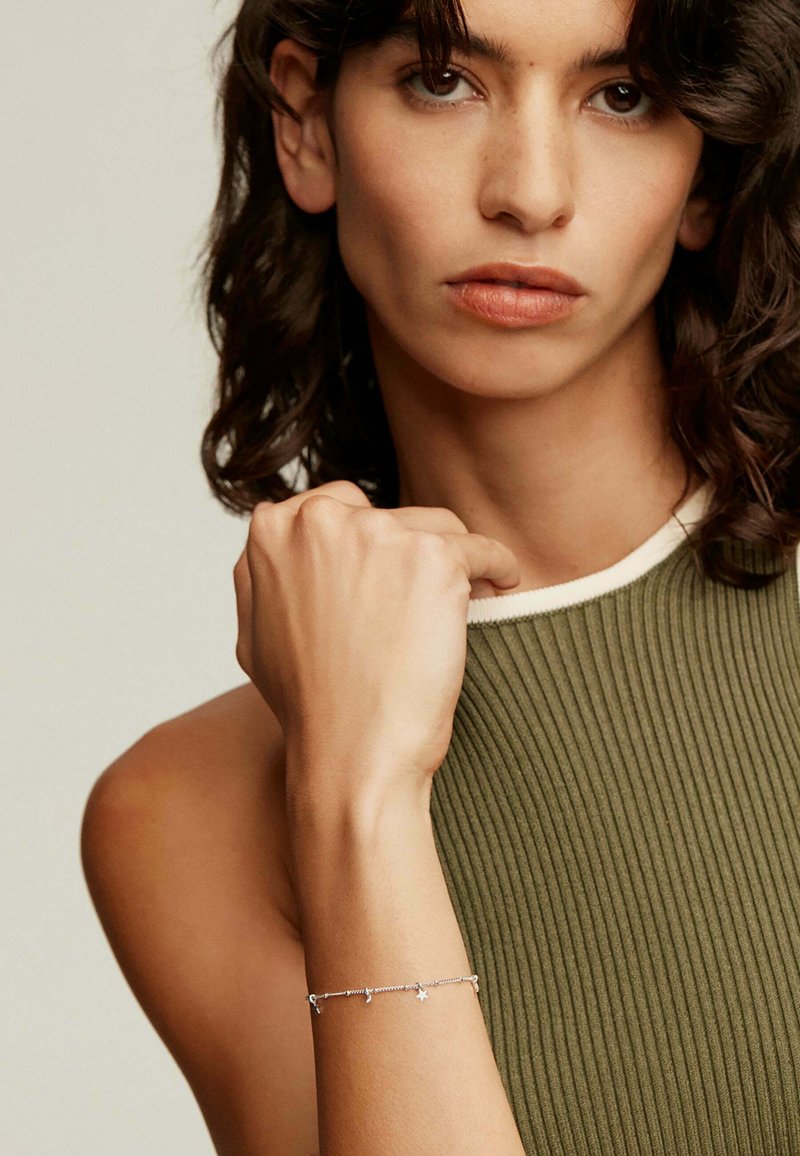 Pulsera de plata con acentos de estrellas llevada en una mano. La blusa de canalé verde del modelo presenta un ribete blanco a lo largo del escote. Textura de piel suave visible.