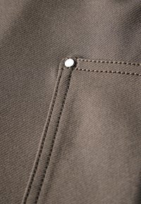 Tissu brun foncé avec une finition texturée, comportant un petit bouton-pression en argent et une couture visible le long du bord de la poche.
