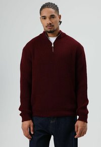 Man staat tegen een grijze achtergrond en draagt een bordeauxrode gebreide trui met een halve ritskraag en donkerblauwe spijkerbroek.