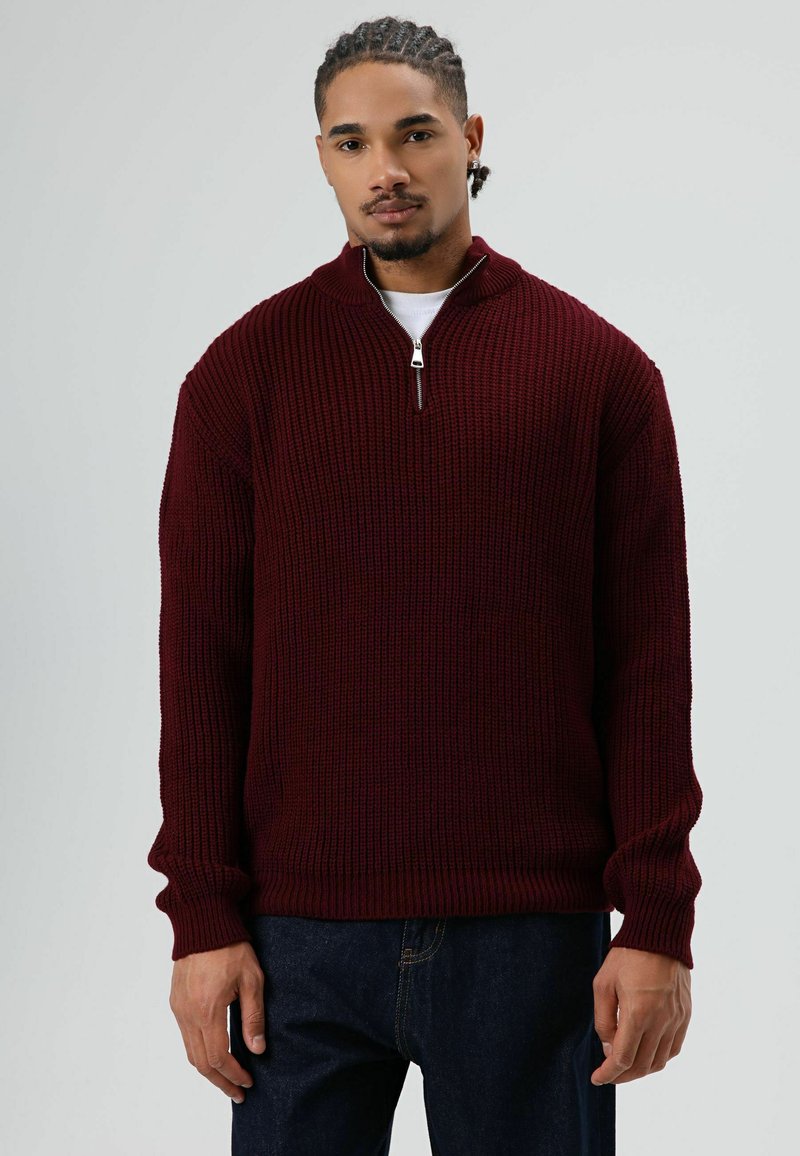 Man staat tegen een grijze achtergrond en draagt een bordeauxrode gebreide trui met een halve ritskraag en donkerblauwe spijkerbroek.