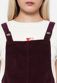 Personne blonde portant une chemise blanche avec deux petits cœurs rouges et une salopette en velours côtelé violet foncé avec des boutons sur les bretelles.