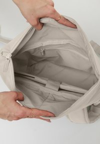 Hands holding open a light beige backpack showing empty main compartment with padded laptop sleeve and inner zippered pocket.