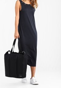 Zwarte stoffen tote bag met dubbele handvatten aan de bovenkant en ritssluiting, met verticale naden en een klein logo-opdruk.