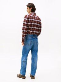 Man standing facing sideways wearing brown plaid shirt, blue jeans, and brown sneakers against a plain white background.