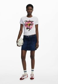 Witte t-shirt met kleurrijke bloemengeheel en zwarte tekst, gecombineerd met een denim mini-jurk, witte tas en rode sportschoenen met gestreepte sokken.