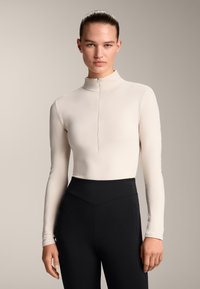 Top lungo beige chiaro con maniche lunghe, colletto alto e chiusura a mezza zip, abbinato a leggings neri a vita alta, tessuto liscio.