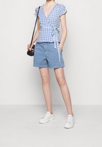 Blusa avvolgente a scacchi blu e bianchi con maniche a volant, abbinata a pantaloni corti azzurri e sneakers bianche, accessoriata con una borsa nera.