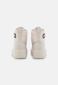 Vaaleanbeige nilkkurit, joissa on pehmustettu muotoilu, teksturoitu pohja ja pienet vetoketjut takana. Logopatchi ulkopuolella.