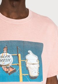 Muž v růžovém tričku s grafikou cedule s kornetem zmrzliny a textem "Cream Castle" na pozadí modré oblohy.