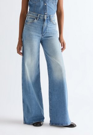 Kobieta ubrana w niebieskie jeansy z wysokim stanem i szerokimi nogawkami z wyblakłymi detalami, w połączeniu z bezrękawnym dżinsowym topem i czarnymi butami na białym tle.