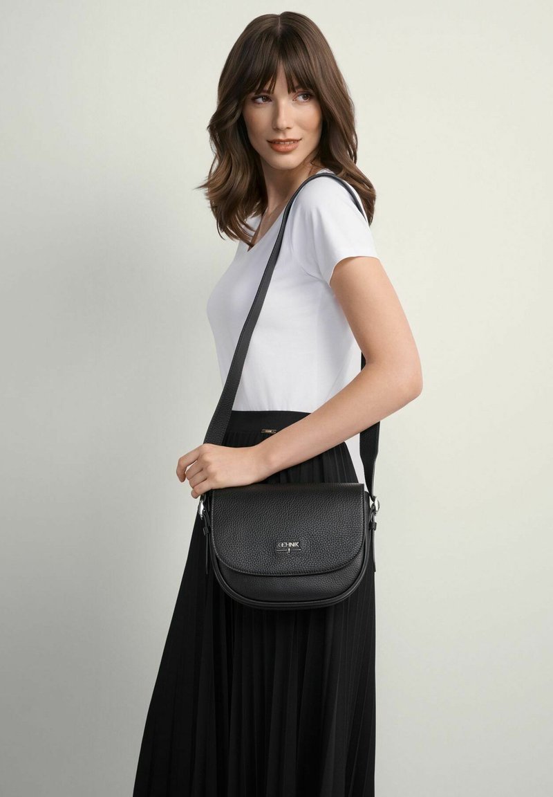 Czarna torba crossbody z teksturowanej skóry, z klapką i regulowanym paskiem. Modelka nosi białą koszulę i czarną spódnicę plisowaną.