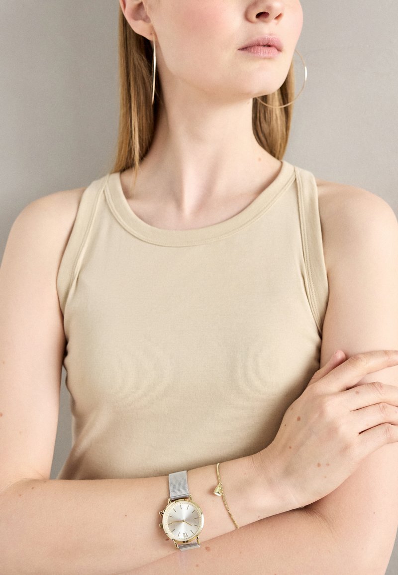 Hõbedane võrkmõõtja valge kellapinnaga ja kuldsete aktsentidega, kantud koos kuldse randmeehtega, millel on väike südameloor. Beez tank top.