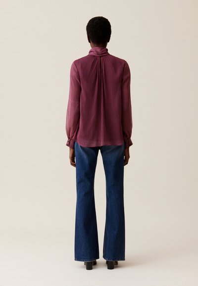 Blusa de manga larga y cuello alto en un burdeos profundo texturizado, combinada con jeans ajustados de cintura alta. Tejido suave con un detalle recogido en la parte posterior.