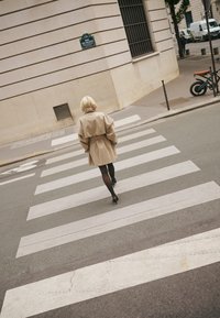 Nő bézs ballonkabátban és fekete harisnyában sétál a gyalogátkelőn a Rue de Valois utcában, ahol parkoló kerékpárok és járművek vannak a városi utcán.