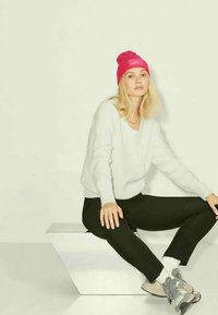Gorro de malha cor-de-rosa, suéter texturizado branco, calças pretas e ténis cinzentos. Sentada sobre um pedestal geométrico branco contra um fundo claro.