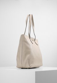 Sac fourre-tout en cuir beige à texture lisse, forme allongée, deux longues anses, et accents noirs subtils sur le dessus. Aucun élément métallique visible.