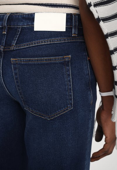 Jeans in denim blu scuro con una sola tasca posteriore caratterizzata da cuciture gialle a contrasto e un'etichetta bianca in vita.