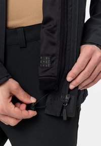 Giacca in tessuto nero con tasca con zip e coulisse regolabile. Etichette visibili per materiali riciclati. Ingrandimento della mano che regola i dettagli.