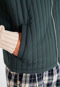 Zeleni jakna s postavom, s patentnim zatvaračem i bočnom džepom, u kombinaciji s kremastim pletenim manžetama i kariranim hlačama, ističe raznolike teksture i boje.