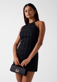 Schwarzes Neckholderkleid aus strukturiertem Stoff mit kontrastierenden Kanten, kombiniert mit einer schwarzen Lederhandtasche mit silberner Logo-Schließe.