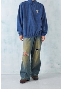 Mörkblå fleecepullover med halvdragkedja, tillsammans med ljusblå slitna baggy jeans med stora hål och fransade kanter.