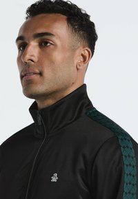 Zwarte zip-up jack met hoge kraag, voorzien van een donkergekleurd groen patroonaccent op de schouder en een klein wit logo op de borst.