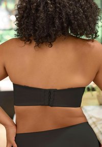 Haut sans manches noir avec une finition lisse, fermeture arrière à crochets avec plusieurs boucles et design minimaliste. Teint de peau visible sur les épaules.