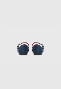 Chaussons en denim avec un dos bas, featuring une bande d'accent rouge et des détails à franges sur les bords. Texture de tissu douce, semelle plate noire.