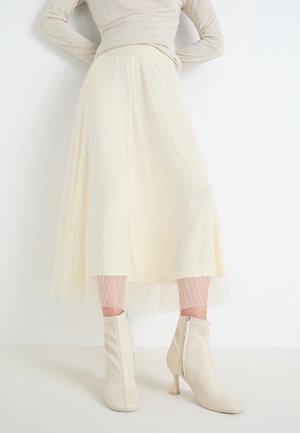 Femme portant une jupe midi plissée crème, un haut beige ajusté à manches longues et des bottines crème à talons, debout contre un mur blanc.