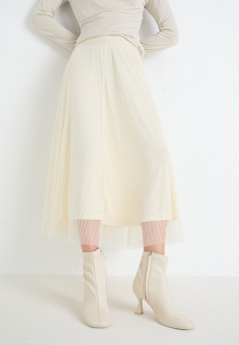 Femme portant une jupe midi plissée crème, un haut beige ajusté à manches longues et des bottines crème à talons, debout contre un mur blanc.