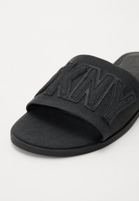 Crne natikače sa slidom s gornjim dijelom od teksturirane tkanine koja ima uzdignuta slova "DKNY". Potplat je ravan i izrađen od gume za bolju prionljivost.