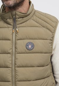 Khakifarbene Pufferweste mit hohem Kragen, gestepptem Design, silbernem Reißverschluss und einem runden Logopatch mit roten, weißen und blauen Akzenten.