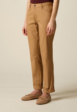 Persona che indossa pantaloni color camoscio risvoltati e mocassini abbinati color camoscio, in piedi davanti a uno sfondo beige uniforme.