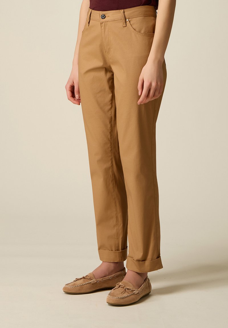 Persona con pantalones color beige enrollados y mocasines a juego, de pie sobre un fondo liso beige.