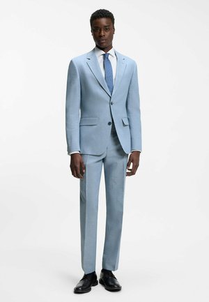 Mann in hellblauem Anzug mit passender Krawatte, weißem Hemd und schwarzen Anzugschuhen, der vor einem einfarbigen weißen Hintergrund steht.
