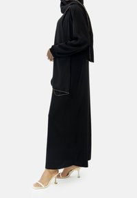 Musta pitkähihaine mekko löysällä istuvuudella ja kootuilla hihoilla. Mukana on yhteensopiva musta huivi, jossa on kultareunus. Yhdistetty valkoisiin remmel sandaaleihin.