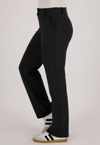 Persoon die zwarte rechte broek en witte sneakers met zwarte strepen draagt, staat met één been gebogen.